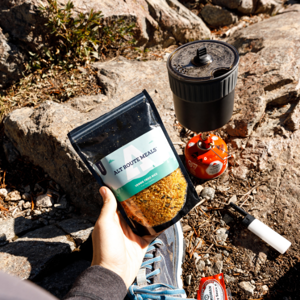 
                  
                    Photo of Alt Route Melas Veggie Fried Rice being held in the palm of a hand. A fuel canister, stove and pot are on the ground in the background.  
                  
                