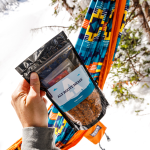 
                  
                    A hand holding the package of Alt Route Meals Thai Curry in the foreground. There is a brightly colored hammock hanging in the trees against a snowy backdrop. 
                  
                