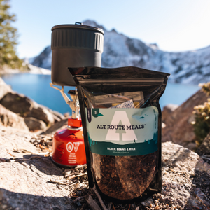 
                  
                    Photo of Alt Route Meals Black Beans and Rice dinner in package sitting on a rock. Blue Lake and Snowy mountains are the background. The fuel canister, stove and pot are pictured directly behind the packaged meal.  
                  
                