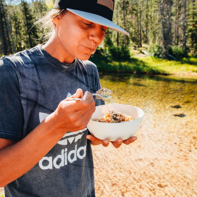 
                  
                    Photo of a woman camper eating the rehydrated Southern Biscuits and Gravy by Alt Route Meals. Standing near water with evergreens in the background. 
                  
                