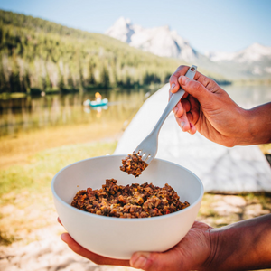 
                  
                    Photo of rehydrated Southwest Hash Brown Scramble by Alt Route Meals. Food is in a white bowl that a hand is holding with a fork coming in to take a bite. Background is a tent set up by a lake that a person can be seen paddling on with mountains in the background.
                  
                
